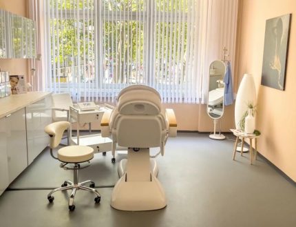 Ein cremefarbener Behandlungsstuhl von hinten betrachtet, steht vor einem Fenster mit weißen Lamellenvorhängen.