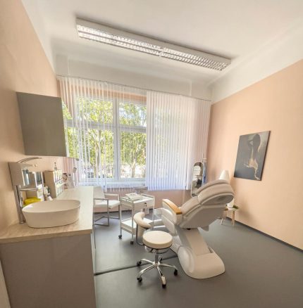 Ein Behandlungszimmer mit apricotfarbenen Wänden, einem Behandlungsstuhl und einem Waschtisch schräg von hinten aufgenommen.